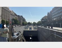 Eingang zur U-Bahn-Station "Unter den Linden" in einer städtischen Umgebung mit breiter Straße, historischen Gebäuden auf beiden Seiten und Radabstellplätzen im Vordergrund. Im Hintergrund sind Bäume und eine Ferneansicht eines Turms sichtbar.