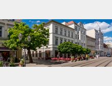 Straßenszene mit historischen Gebäuden in hellen Fassaden, einem Straßencafé mit roten Stühlen unter Bäumen und Straßenbahnschienen im Vordergrund. Im Hintergrund sind blauer Himmel und vereinzelte weiße Wolken sichtbar.