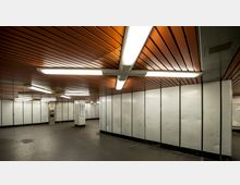 U-Bahn-Station mit einer Decke aus orangefarbenem Holz und hellem Neonlicht. Die Wände sind mit grauen Platten verkleidet, und in einer Ecke befindet sich ein Münztelefon neben einem Pfeiler.