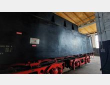 Ein großer schwarzer Wassertank eines historischen Schienenfahrzeugs in einer Halle mit Holzdachkonstruktion und hohen Fenstern; rote Fahrwerkskomponenten und Hinweisschilder wie „Vorsicht! Kein Trinkwasser“ sind sichtbar.