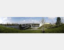 Weitläufige Grünanlage mit jungen Bäumen und Spazierwegen, im Hintergrund eine große, moderne Straßenüberführung mit mehreren Fahrspuren und Betonpfeilern, umgeben von städtischer Infrastruktur und vereinzelten Gebäuden.