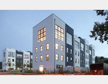 Moderne Wohngebäude mit mehreren Stockwerken in klarer, kubischer Bauweise. Die Fassade ist in Weiß und Dunkelgrau gehalten, große Fenster prägen das Design, und einige Fenster sind erleuchtet; im Vordergrund befinden sich eine Straße und parkende Autos.