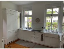 Helle Küche mit weißen Schrankfronten im Landhausstil, einer Arbeitsplatte aus Marmoroptik und zwei großen Fenstern, die Blick auf einen Garten bieten. Die Einrichtung umfasst eine Spüle, Elektrokochfeld und Hängelampen, mit Holzboden und dezenten Dekorationen.