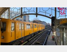 Gelber U-Bahn-Waggon der Berliner Verkehrsbetriebe (BVG) steht an einer oberirdischen Station mit Glasfenstern und geschwungenen Metallbögen. Im Hintergrund sind Gebäude, ein Signallicht und Teile der Gleise zu sehen.