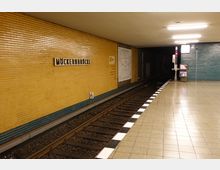 U-Bahn-Station "Möckernbrücke" mit gelben gefliesten Wänden und einem Wandplan. Auf der rechten Seite befindet sich ein Bahnsteig mit hellen Fliesen, die zu den Gleisen führen, während die Wand auf der linken Seite mit dem Stationsnamen beschriftet ist.