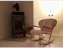 Gemütliche Ecke mit einem modernen Kaminofen, der auf einer Glasplatte steht, darunter gestapeltes Brennholz. Daneben ein Schaukelstuhl mit gewebter Sitzfläche und einer weißen Fellauflage, im Hintergrund ein dezentes, warmes Licht und eine geschlossene weiße Schranktür.