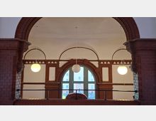 Innenansicht eines Flurs mit großen Backsteinbögen und weiß verputzten Gewölbedecken, beleuchtet von drei runden Hängelampen. In der Mitte befindet sich ein hohes Rundbogenfenster mit Backsteinrahmung und einem Geländer davor; draußen sind Fassaden gegenüberliegender Gebäude zu sehen.