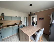 Moderne Küche mit hellblauen Schränken, Holzarbeitsflächen und einem zentralen, hölzernen Küchentisch mit zwei gepolsterten Barhockern. Im Hintergrund eine geöffnete Tür zu einem angrenzenden Raum mit Dekorationsobjekten und Pflanzen.