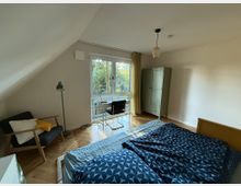 Ein helles Schlafzimmer mit Dachschräge, einem großen Fenster mit Blick auf Bäume, einem Bett mit blauer Bettdecke, einem Schreibtisch mit Stuhl, einem Sessel und einem grünen Kleiderschrank. Der Boden ist aus Holzparkett, und eine Pendelleuchte hängt an der Decke.