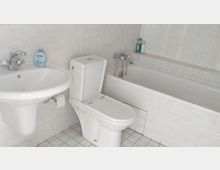 Ein Badezimmer mit weiß gefliesten Wänden und Boden. Es zeigt ein Waschbecken mit Seifenspender, eine Toilette und eine Badewanne mit Duscharmatur.