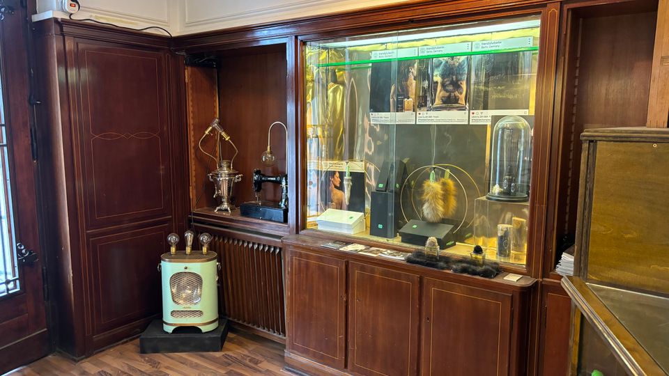 Innenraum mit dunkler Holzvertäfelung und eingebauten Vitrinen, in denen verschiedene Ausstellungsobjekte und Poster hinter Glas präsentiert werden. Vor der Vitrine stehen ein kleines Retro-Heizgerät und auf einer Nische sind dekorative Geräte wie eine alte Nähmaschine und ein Samowar zu sehen.