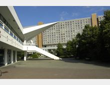 Ein moderner Platz mit einem weißen Gebäude, das durch markante geneigte Stützen und eine gerade Außentreppe geprägt ist. Im Hintergrund ist ein mehrstöckiges Wohn- oder Bürogebäude mit vielen Fenstern zu sehen, flankiert von Bäumen auf der rechten Seite.