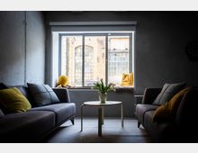 Modernes Wohnzimmer im Industriestil mit zwei grauen Sofas, gelben Kissen und einem runden Couchtisch mit Vase in der Mitte. Großes Fenster mit breiter Fensterbank und Kissen, Blick auf ein Backsteingebäude mit hohen Sprossenfenstern.