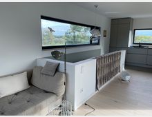 Modernes Wohnzimmer mit hellem Holzfußboden, einem grauen Sofa und einer Stehlampe. Im Hintergrund befinden sich breite, rechteckige Fenster mit Ausblick auf Bäume und eine moderne Einbauküche in Grau.