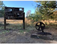 Eine Holz- und Metallkonstruktion steht auf einer Wiese inmitten eines Waldes; links eine Insektennisthilfe aus Holzscheiten und Ästen, rechts ein verrostetes, metallisches Schwungrad auf einem Holzblock. Im Hintergrund sind Kiefern und offenes Grasland sichtbar.
