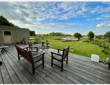 Holzterrasse mit Gartenmöbeln wie Holzbänken und einem runden Tisch, dahinter ein großer, gepflegter Rasen mit Blumenbeeten, Bäumen und weitläufiger Landschaft unter einem Himmel mit leichter Bewölkung. Links steht ein kleiner Holzschuppen.