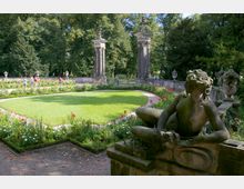 Historischer Garten mit gepflegtem Rasen und Blumenbeeten, umgeben von einer steinernen Balustrade. Im Vordergrund eine Skulptur einer liegenden Figur, im Hintergrund zwei dekorative Säulen und dichte Bäume.