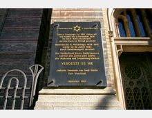 Gedenktafel an der Außenwand einer Synagoge aus Backstein mit rundbogigem Fenster und verziertem Schmiedeeisengitter. Die Tafel erinnert an die Zerstörung der Synagoge während der Pogromnacht 1938 und des Zweiten Weltkriegs.