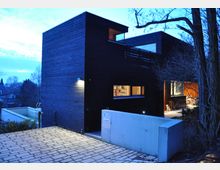 Ein modernes Wohnhaus aus dunklem Holz mit kubischen Formen, aufgenommen bei Dämmerung. Vor dem Gebäude befindet sich eine gepflasterte Einfahrt, eine Betonmauer grenzt den Eingangsbereich ab, und im Hintergrund sind Bäume und ein Pool erkennbar.