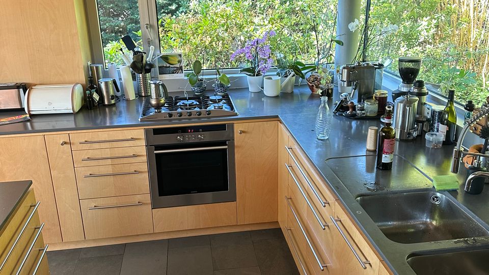 Moderne U-förmige Küche mit hellen Holzschränken, dunkelgrauen Bodenfliesen und Edelstahl-Arbeitsflächen mit integriertem Herd, Backofen und Doppelspüle. Entlang der Arbeitsplatte verläuft ein breites Fensterband mit Blick ins Grüne und einigen Pflanzen auf der Fensterbank.