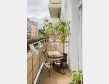 Ein schmaler Balkon mit einem Korbstuhl, auf dem ein gemustertes Kissen liegt, umgeben von mehreren Topfpflanzen. Die Brüstung ist mit einer Lichterkette geschmückt, und eine Katze liegt auf einem beige gemusterten Teppich. Im Hintergrund sind Wohnhäuser und Bäume zu sehen.