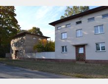 Zwei einfache Wohnhäuser mit Flachdächern in einer ruhigen Wohngegend. Das linke Gebäude hat eine graue, unverputzte Fassade, während das rechte Gebäude verputzt ist; im Vordergrund ist eine kleine Rasenfläche und eine Straße sichtbar.