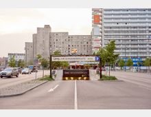 Einfahrt zum Parkhaus "Q-Park am Alexanderplatz" in einer belebten urbanen Umgebung mit mehrspuriger Straße, flankiert von hohen Gebäuden im modernen Stil. Im Hintergrund sind Wohn- und Bürogebäude sowie Werbetafeln zu sehen.