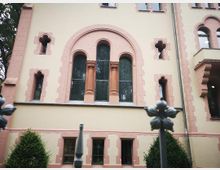 Fassade eines historischen Gebäudes mit gelblicher Putzwand und rosa Sandsteinelementen, darunter Spitzbogenfenster, kleine gotische Fensterformen sowie Dekorsäulen. Im Vordergrund ein schmiedeeiserner Zaun mit floralen Ornamenten und ein Busch.