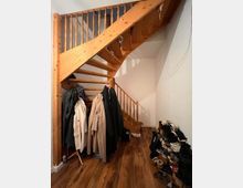 Holztreppe mit einem Geländer aus senkrechten Holzstäben in einem Eingangsbereich; darunter hängen Mäntel auf einer Garderobe, und rechts daneben stehen mehrere Paar Schuhe auf einem Schuhregal und auf dem Boden. Der Raum hat Holzfußboden und weiße Wände.