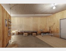 Minimalistisch eingerichteter Innenraum mit Wänden aus hellem Sperrholz und einer Betondecke. Der Raum enthält eine Sitzbank mit Kissen, geflochtene Hocker, ein Regal mit Zeitschriften und einen kleinen Holztisch mit Dekorationszweigen.