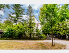 Ein weißes Haus mit Satteldach, teilweise verdeckt durch dichte grüne Bäume und Sträucher, umgeben von einem Metallzaun. Im Vordergrund befindet sich ein Gehweg, daneben eine Wiese mit vereinzelten Bäumen.