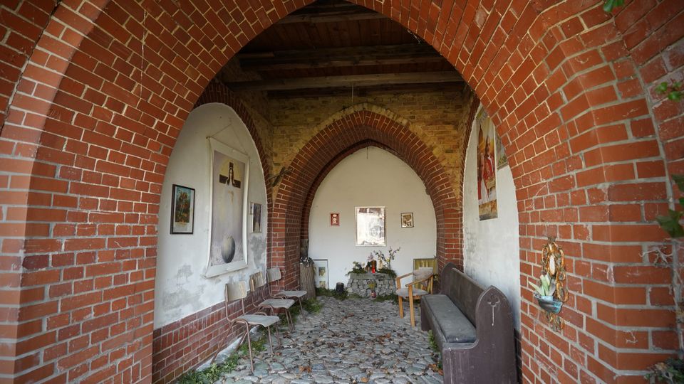 Überdachter Gang oder kleine Kapelle mit spitzbogigen Backsteinarkaden und Kopfsteinpflasterboden, an den weiß verputzten Wänden hängen religiöse Bilder. Entlang der Seiten stehen einfache Stühle und eine Holzbank; am Ende befindet sich ein kleiner Altar mit Blumen und Kerzen.