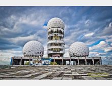 Verlassenes Radom-Gelände mit drei großen weißen Kugelkuppeln und einem zentralen zylindrischen Turm, dessen Fenster teils zerstört sind. Davor liegt eine weitläufige, rissige Betonfläche, die Gebäude sind mit Graffiti bedeckt, im Hintergrund ein wolkiger Himmel.