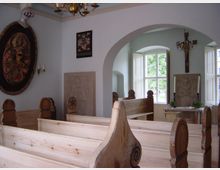 Kleine, helle Kapelle mit Holzbänken, einem Kreuz an der Wand und reich dekorierten Wandschnitzereien. Durch die großen Fenster fällt Tageslicht ein, und ein Altar mit Kerzen und Blumenschmuck steht im Hintergrund.