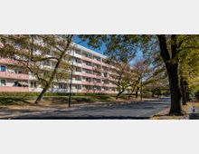 Mehrstöckiges Wohngebäude mit rosafarbenen und weißen Balkonen, umgeben von Bäumen mit herbstlichem Laub. Im Vordergrund verläuft eine asphaltierte Straße, daneben befinden sich Gehwege und Rasenflächen.