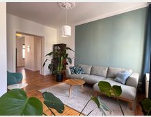 Wohnzimmer mit hellgrauem Sofa vor einer mintgrünen Wand, dekoriert mit Kissen. Holzboden, ein runder Couchtisch, ein Teppich und Zimmerpflanzen prägen den Raum. Im Hintergrund ist ein Türdurchgang zu einem weiteren Raum sichtbar.