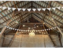 Innenansicht einer rustikalen Scheune mit Holzbalken und Ziegeldach. An der Decke hängen weiße Wimpelgirlanden, Lichterketten und ein Kronleuchter, darunter sind unverputzte Ziegelwände sichtbar.