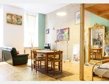 Wohnzimmer mit Holzboden, Essbereich mit Holztisch und Stühlen, einer grünen Ledercouch und einer hellen Ziegelwand mit Kunstwerken. Ein großer vergoldeter Spiegel lehnt an der Wand, und rechts ist ein Teil der Küche sichtbar.