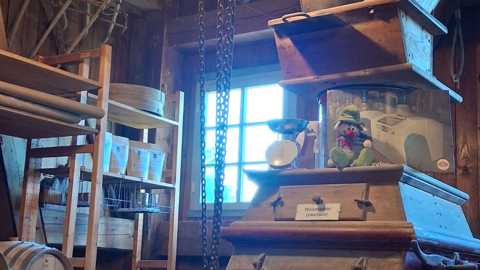 Innenraum einer rustikalen Werkstatt oder Mühle mit Holzbalken, Fenster und hängendem Kettenzug unter der Decke. Im Vordergrund steht eine große, alte Metallmaschine, umgeben von Regalen mit Werkzeugen und Gegenständen sowie einem Holzfass.