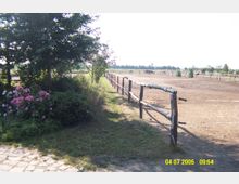 Reitplatz mit einem Holzzaun, umgeben von Bäumen, Blumen und Wiese. Im Hintergrund sind Weiden und einige Pferde zu erkennen, die in einer ländlichen Umgebung stehen.