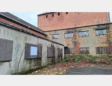 Hinterhof eines verfallenen Industrie- oder Lagergebäudekomplexes mit bröckelndem Putz, freiliegendem Ziegelmauerwerk und vergitterten Fenstern. Vor den Gebäudewänden liegen überwucherte Sträucher und Laub entlang einer teils beschädigten Asphaltfläche.