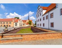 Historische Schlossanlage mit weiß verputztem Hauptgebäude und roten Ziegeldächern, umgeben von einer Ziegelsteinmauer und einem gepflasterten Vorplatz; im Hintergrund eine Kirche mit spitzem Turm.