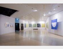 Innenraum einer modernen Kunstgalerie mit hell beleuchtetem Holzfußboden und weißen Wänden. An den Wänden hängen bunte Gemälde und ein großes blau-weißes Banner, in der Mitte steht eine schlichte Holzbank.