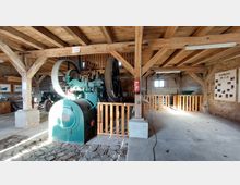 Innenansicht eines historischen Maschinenraums mit einer großen, grünen Industriemaschine in der Mitte. Der Raum verfügt über Holzbalkendecken, Ziegelwände und Holzgeländer, mit Infotafeln an den Wänden sowie einem kleinen Fenster, das Tageslicht hereinlässt.