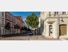 Straßenszene in einer Wohngegend mit historischen Gebäuden im Stil der Gründerzeit, links ein rotes Backsteinhaus mit weißen Verzierungen, rechts ein hell verputztes Eckgebäude mit geschwungenen Fassadenelementen. Gepflasterte Fahrbahn mit parkenden Autos, ein einzelner Baum und Verkehrsschilder mit Geschwindigkeitsbegrenzung „30 Zone" sowie Parkverbot prägen den Straßenabschnitt.