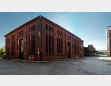 Backsteingebäude mit industrieller Architektur, charakterisiert durch hohe, schmale Fenster und vertikale Verzierungen an der Fassade. Das Gebäude steht auf einem gepflasterten Platz unter freiem Himmel, umgeben von weiteren Industrieanlagen.