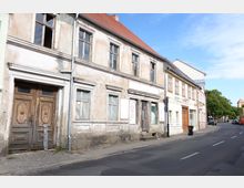 Straßenszene mit einem alten Gebäude mit bröckelnder Fassade, teils kaputten Fenstern und einer großen Holztür. Daneben ein renoviertes Gebäude mit Geschäften, einer schmalen Straße und vereinzelten Bäumen im Hintergrund.
