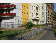 Straßenszene mit mehrstöckigen Wohnhäusern in verschiedenen Farben, darunter eine gelbe und eine beige Fassade. Im Vordergrund befindet sich ein gepflasterter Gehweg mit einem Metallgeländer, Wiesenflächen und einige Büsche, während im Hintergrund weitere Gebäude und ein parkendes Auto zu sehen sind.