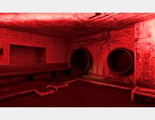 Ein unterirdischer Raum mit roter Beleuchtung, Betonwänden und sichtbaren runden Belüftungsöffnungen. Eine lange Sitzbank ist entlang der linken Wand angebracht, und der Boden hat Stufen und erhöhte Bereiche.