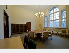 Ein Konferenzraum mit beigen Wänden, einem runden Holztisch und mehreren Polsterstühlen. Große, halbrunde Fenster lassen Tageslicht herein, daneben befindet sich ein Heizkörper; im Hintergrund stehen ein Wandschrank und eine Tür aus dunklem Holz.